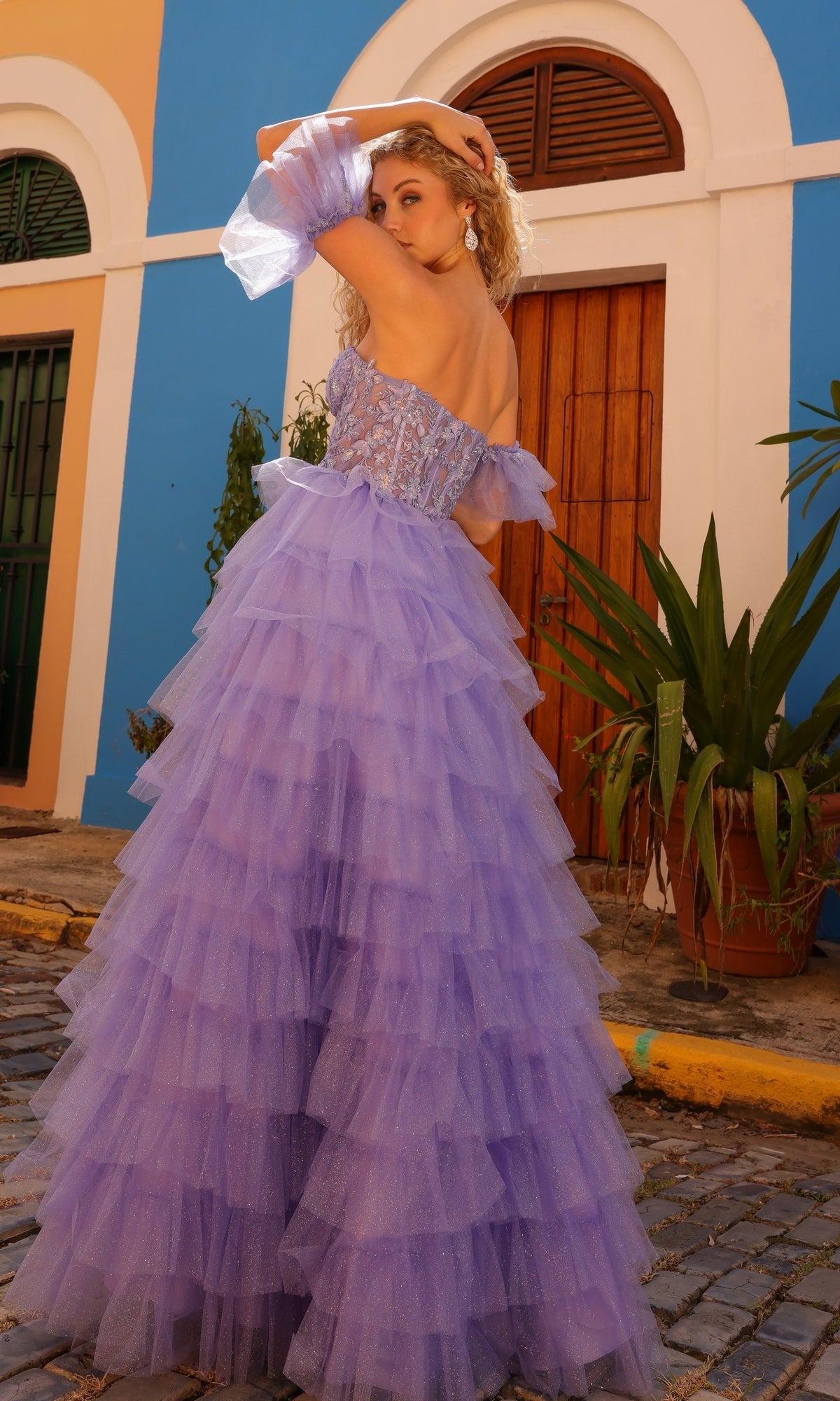 A woman stands on a cobblestone street with colorful buildings and potted plants, wearing the Puff-Sleeve Strapless Long Ruffle Prom Dress T1338 in lavender, featuring delicate embroidered flowers and layered ruffles.